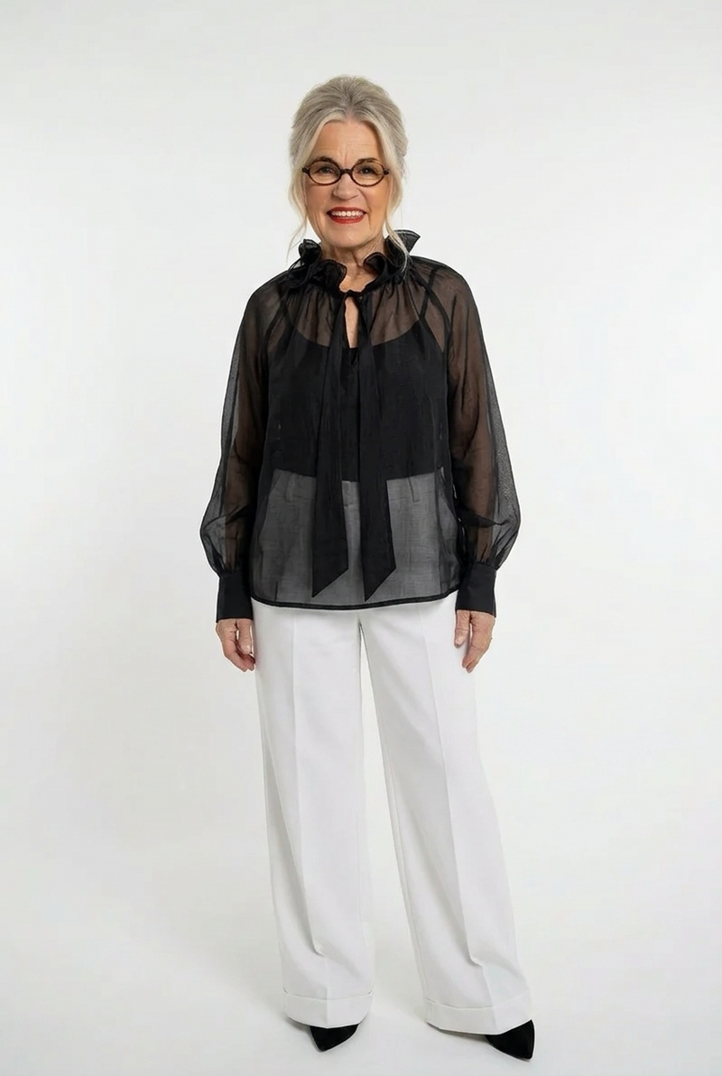 musta läpikuultava sifonkipusero, jossa pitkät hihat, frillakaulus ja nauhat pääntiellä. Naisen päällä valkoisten housujen kanssa.