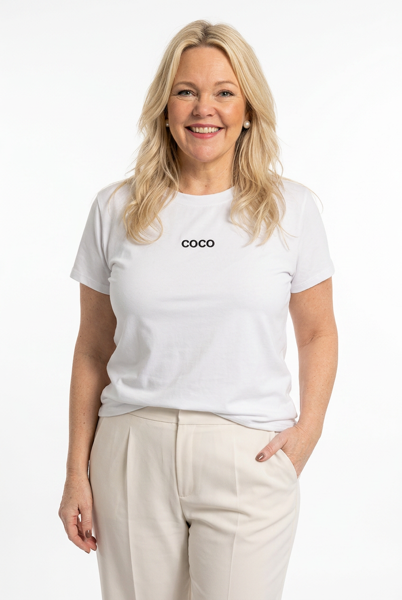 naisen päällä valkoinen t-paita lyhyellä hihalla ja o-pääntiellä, jossa pieni tumma coco logo edessä.